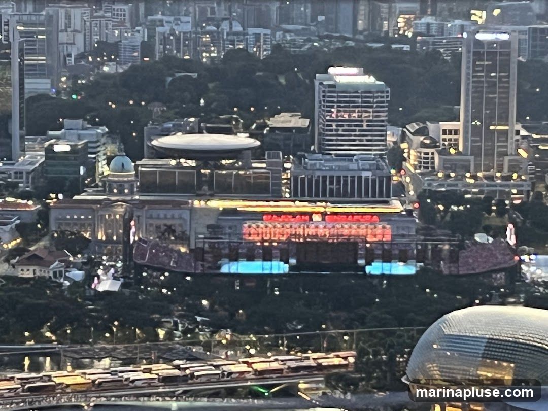 An aerial view of a city at dusk, showcasing a cluster of buildings. A large, prominent building in the center is illuminated with red and blue lights, and features large text that appears to be in a foreign language. Surrounding it are other structures of varying heights, some with illuminated windows. In the foreground, a domed structure with a patterned surface reflects the ambient light. The background consists of a dense urban landscape with numerous high-rise buildings and a dark, tree-covered hill.