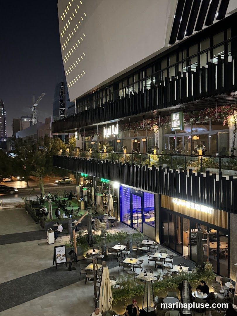 An outdoor dining area with multiple tables and chairs is set up in front of a modern building at night. The building has large windows and balconies adorned with plants and flowers. Several people are seated at the tables, enjoying their meals or drinks. The area is well-lit, creating a warm and inviting atmosphere.