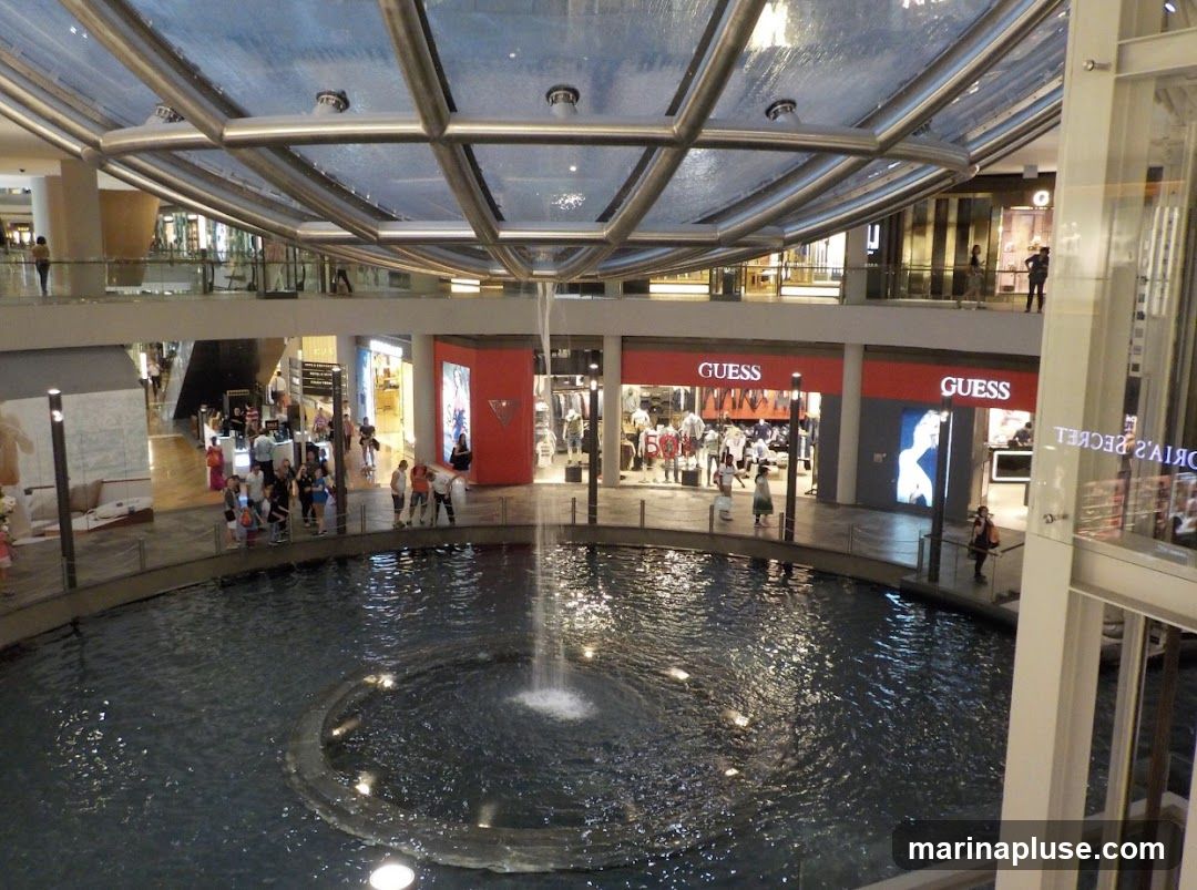 An indoor shopping mall features a large circular fountain in the center, with water cascading from above. Surrounding the fountain are multiple levels of shops, including a prominent GUESS store with its signage visible. People are seen walking around the fountain and browsing in the shops. The architecture includes a modern glass ceiling and balconies overlooking the central area.