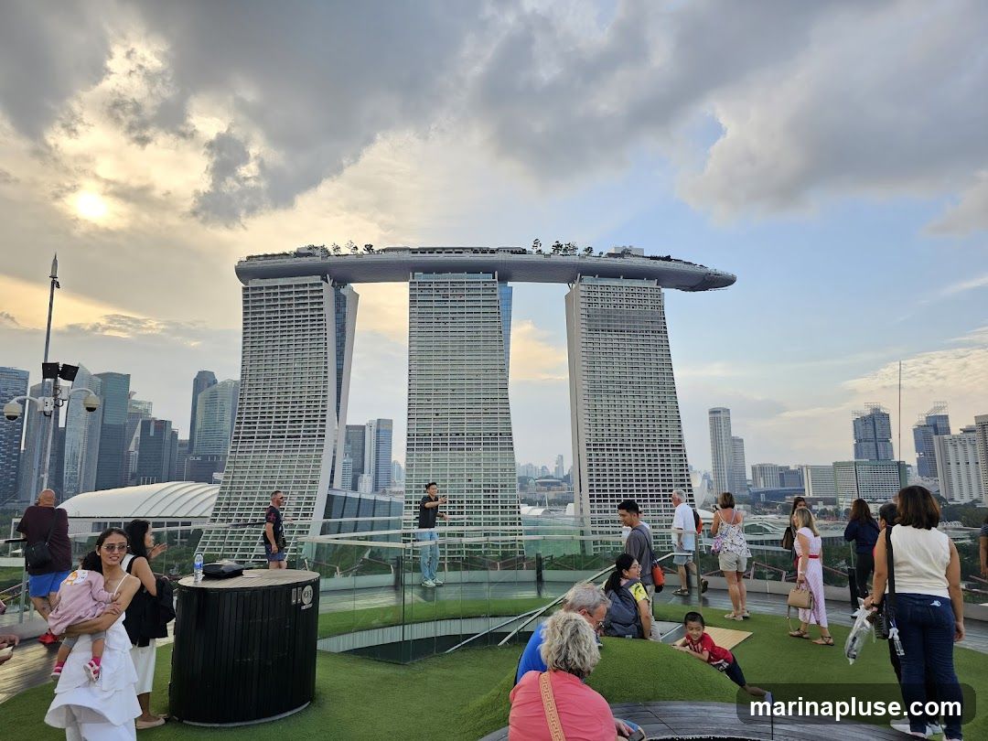A group of people are gathered on a rooftop observation deck, with the iconic Marina Bay Sands hotel in the background. The sky is partly cloudy with the sun setting. The people are casually dressed and appear to be tourists taking in the cityscape.