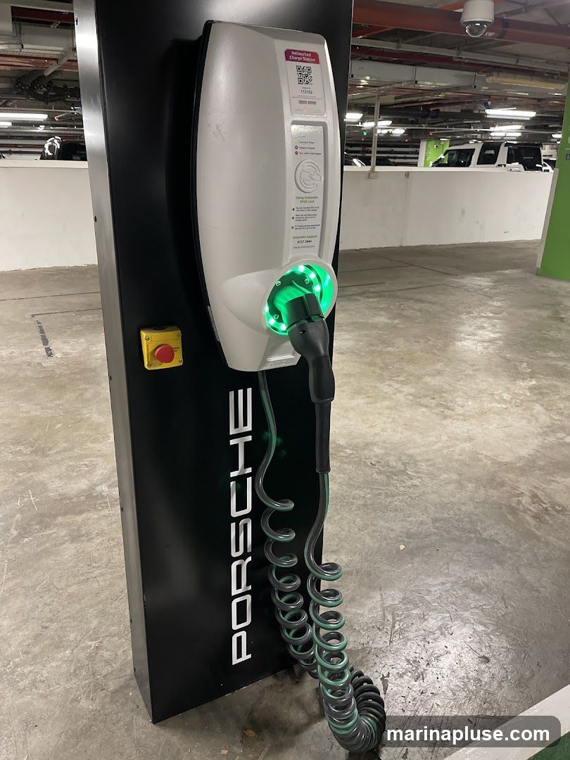 A black charging station with the Porsche logo vertically displayed on its side. A white charging unit is attached, with a green charging cable and connector plugged in and illuminated. The station is located in a parking garage, with other vehicles visible in the background.