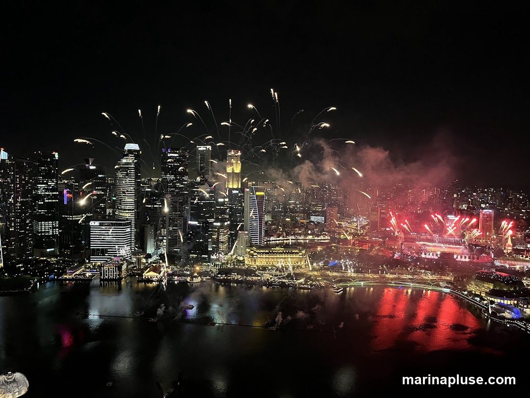 A panoramic view of a city skyline at night, illuminated by a spectacular fireworks display. Numerous skyscrapers are lit up, reflecting on the dark water in the foreground. The fireworks are bursting in various colors, predominantly white and red, with trails of smoke adding to the dramatic effect. The scene captures a festive and vibrant atmosphere.