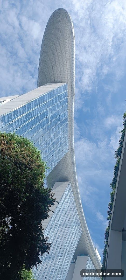 A low-angle shot of a modern skyscraper with a distinctive curved, boat-like roof structure. The building features a facade of glass panels and geometric patterns on the roof. Lush green trees are visible in the foreground, and a bright blue sky with scattered clouds forms the background.