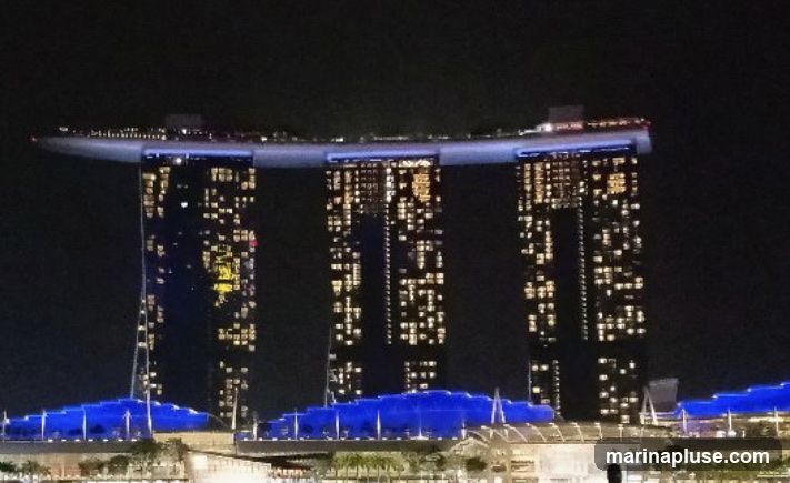 A nighttime view of the Marina Bay Sands hotel in Singapore, featuring its iconic three towers connected by a rooftop deck. The building is illuminated with many lit windows, and the rooftop area is lit with blue lights. The lower levels of the complex are also visible, with lights and architectural details.