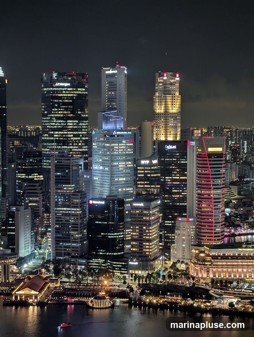 A nighttime aerial view of a dense cityscape with numerous illuminated skyscrapers and a waterfront area. The buildings display a variety of architectural styles and lighting, with some featuring prominent company logos such as J.P. Morgan and Singapore Land. The waterfront is lined with buildings and structures, some of which are also lit up, and a body of water reflects the lights. A small red boat is visible on the water.