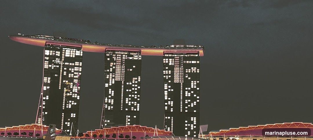 A nighttime view of the iconic Marina Bay Sands hotel in Singapore, with its three towers illuminated by numerous lights. The distinctive boat-shaped Sands SkyPark is visible at the top, glowing with warm orange and pink hues. The lower structures of the complex are also lit, creating a vibrant urban landscape against the dark sky.