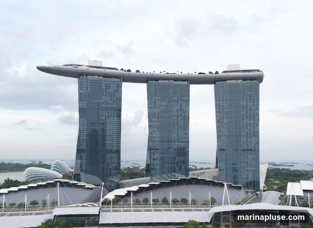A wide shot of the Marina Bay Sands hotel in Singapore, featuring its iconic three towers connected by a sky park. The hotel is set against a cloudy sky, with a body of water and other buildings visible in the background.