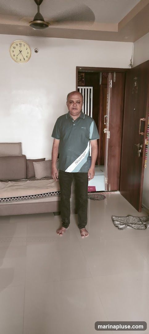 A man stands in a room, facing the camera. He is wearing a dark green polo shirt with a white and light blue stripe on the side, and dark pants. He is barefoot. To his left is a sofa with several cushions. Behind him, a doorway is visible, leading to another room. A clock is mounted on the wall above the sofa. The floor is tiled.