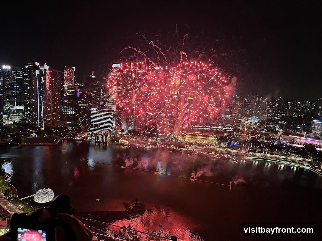 A vibrant fireworks display illuminates the night sky over a densely populated city skyline. Red fireworks explode in large bursts, casting a warm glow on the surrounding buildings and the body of water below. The cityscape features numerous skyscrapers and illuminated structures, reflecting the festive atmosphere. In the foreground, a person is visible holding a camera, seemingly capturing the spectacle.