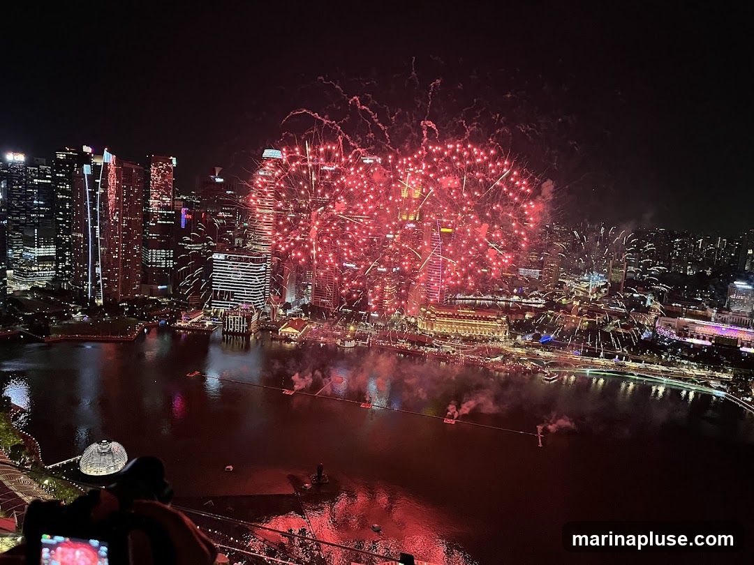 A vibrant fireworks display illuminates the night sky over a densely populated city skyline. Red fireworks explode in large bursts, casting a warm glow on the surrounding buildings and the body of water below. The cityscape features numerous skyscrapers and illuminated structures, reflecting the festive atmosphere. In the foreground, a person is visible holding a camera, seemingly capturing the spectacle.