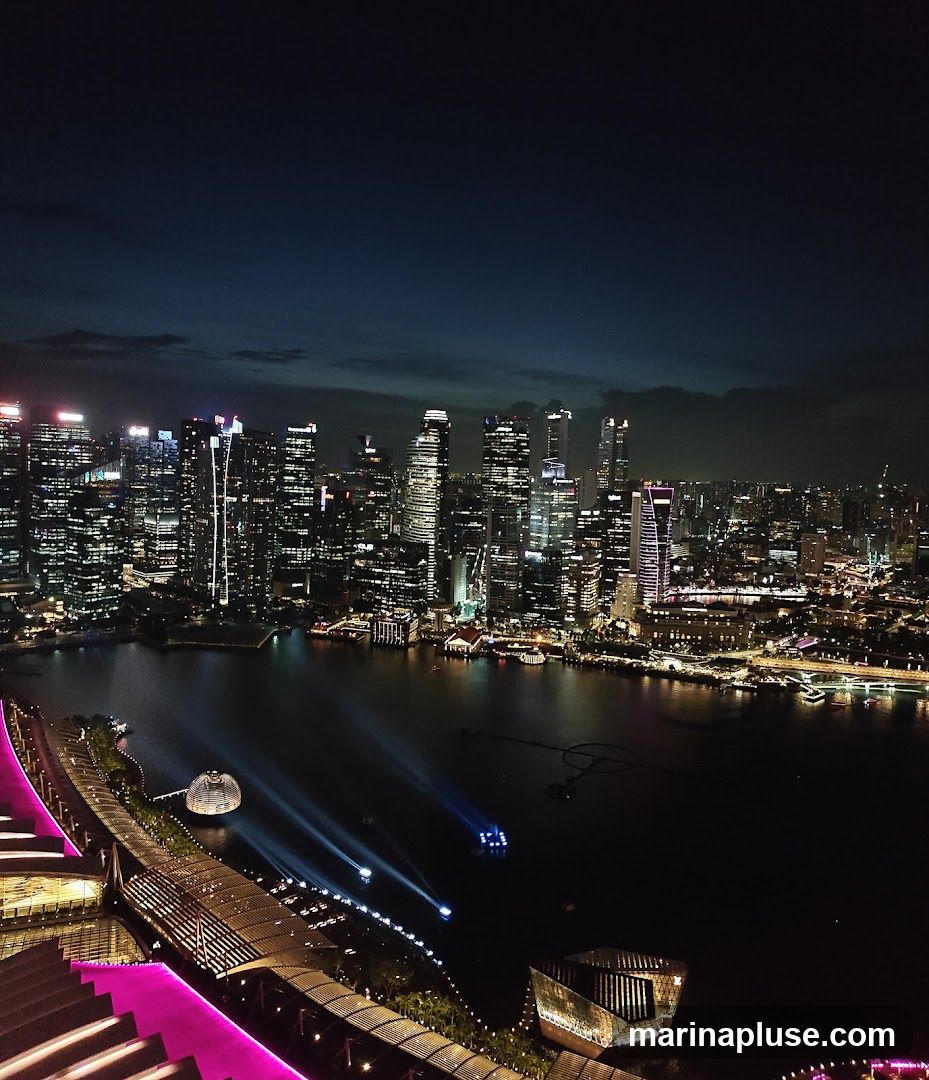 A panoramic view of a city skyline at night, illuminated by countless lights from skyscrapers. A dark body of water, likely a river or bay, separates the foreground from the distant buildings. Several boats are visible on the water, some emitting beams of light. In the foreground, a structure with vibrant pink lighting and a tiered design suggests a rooftop or elevated walkway, possibly part of a resort or entertainment venue. The sky above is a deep, dark blue with some scattered clouds.