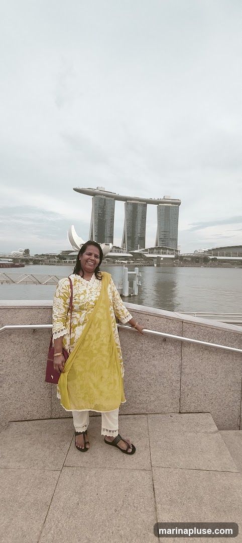 A woman smiles at the camera while standing on a stone walkway. Behind her, the iconic Marina Bay Sands hotel in Singapore is visible across a body of water. The sky is overcast.