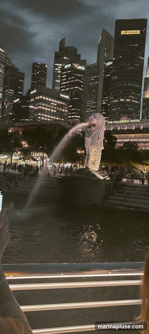 A nighttime view of the Merlion fountain in Singapore, with the city skyline in the background. The Merlion statue is spouting water into the river. Several buildings are visible, including The Fullerton Hotel, and buildings with signs for MYP and wework. People are gathered around the fountain area.