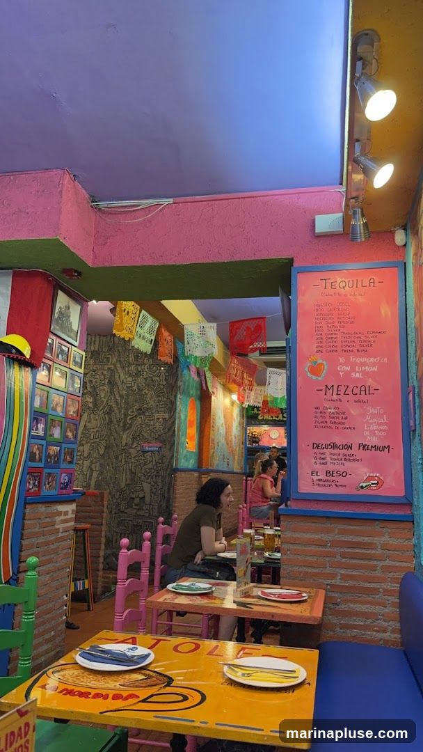 A pink menu board displays selections of tequila and mezcal, along with a premium tasting option and a cocktail called 'El Beso'. The menu is set against a backdrop of colorful decor and brickwork.