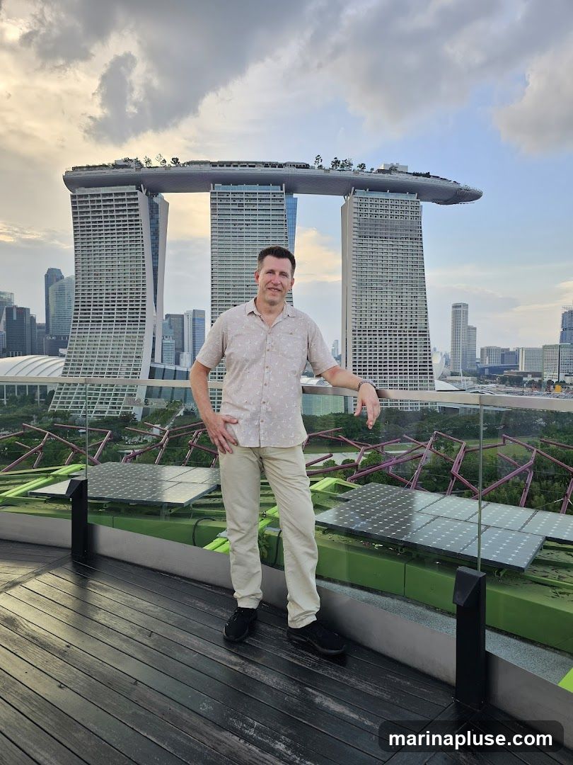 A man stands on a wooden deck, posing for a photo with the iconic Marina Bay Sands hotel in Singapore in the background. The sky is cloudy, with the sun setting or rising. In the foreground, there are solar panels and greenery.
