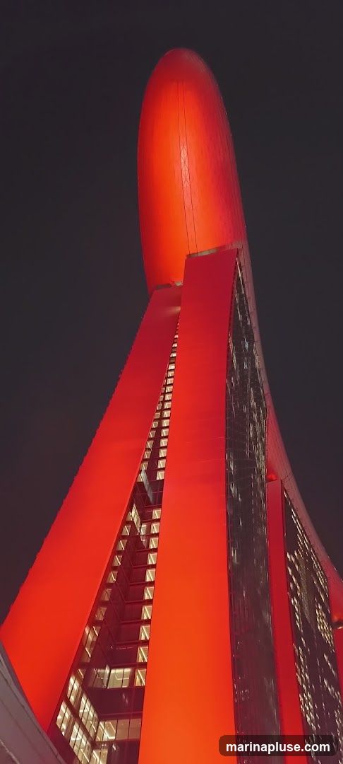 A striking, modern building with a distinctive red exterior, illuminated against a dark night sky. The structure features a large, bulbous red element at the top, with sweeping red panels that create dramatic angles and reveal glimpses of illuminated windows within the building's facade. The architecture is contemporary and eye-catching.