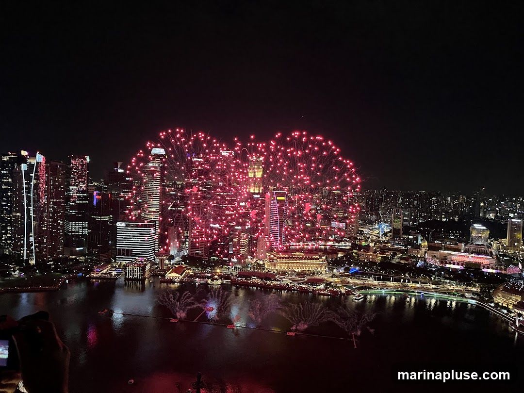 A vibrant fireworks display illuminates the night sky above a densely packed city skyline. Red fireworks burst in large, heart-shaped patterns, casting a warm glow on the surrounding skyscrapers and buildings. The city lights twinkle below, reflecting on the dark water in the foreground. Several boats are visible on the water, possibly part of the event. A hand holding a camera is visible in the lower left corner, capturing the spectacle.
