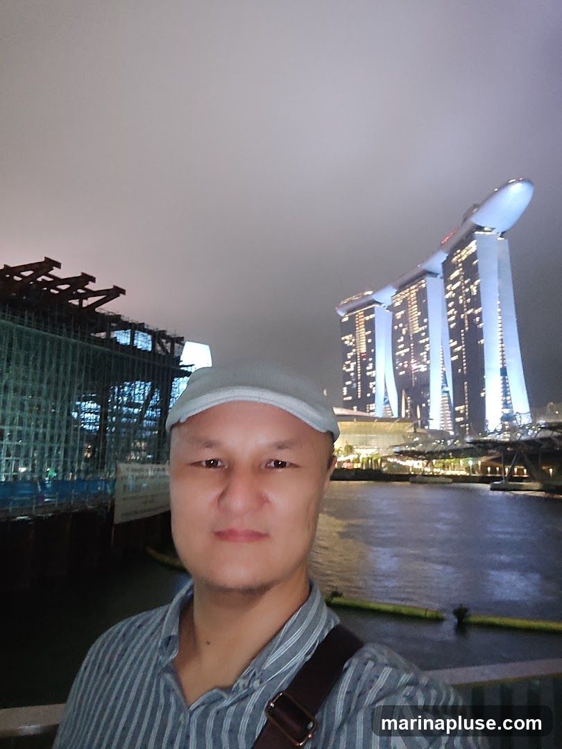 A man in a striped shirt and a cap takes a selfie at night. In the background, the iconic Marina Bay Sands hotel in Singapore is illuminated, along with a construction site on the left. The water in front reflects the city lights.