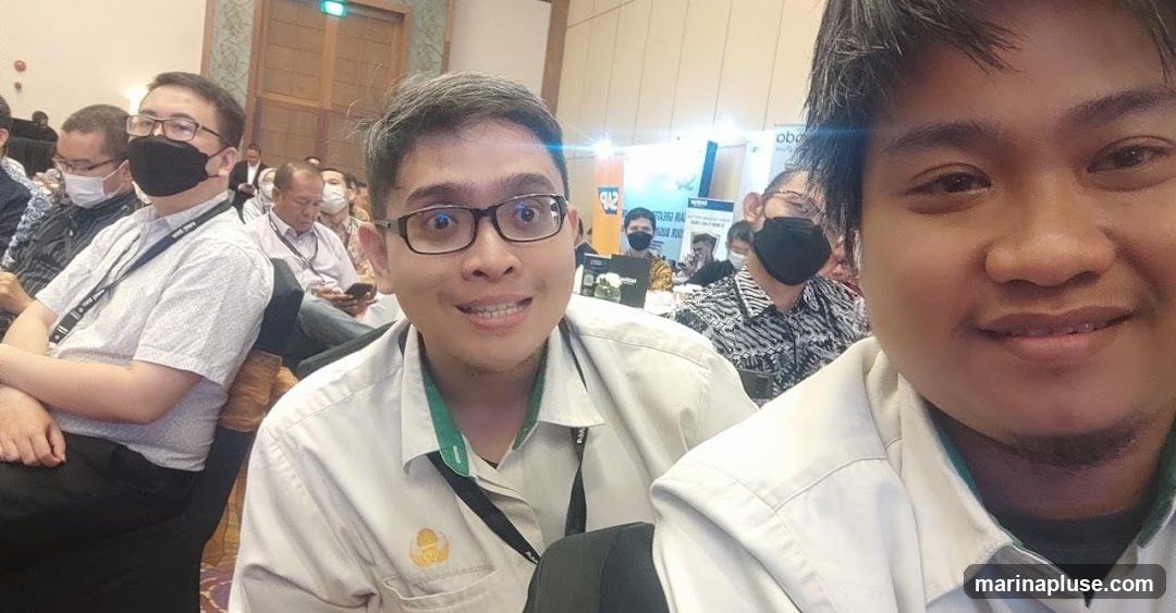 Two men in the foreground are taking a selfie at a conference or event. They are wearing light-colored collared shirts and lanyards. In the background, other attendees are seated, some wearing masks. There are also banners and displays visible behind the seated individuals.