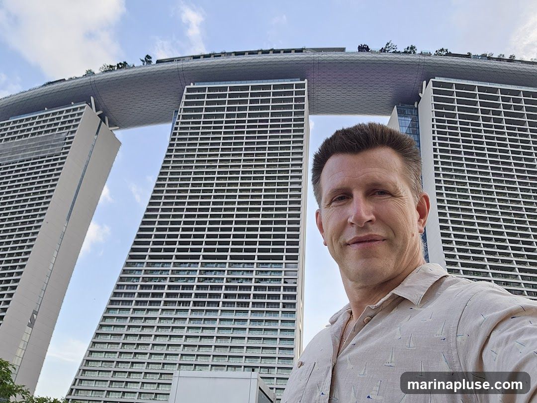 A man takes a selfie in front of the iconic Marina Bay Sands hotel in Singapore. The hotel's distinctive three towers, connected by a large rooftop deck, are visible behind him under a partly cloudy sky. The man is wearing a light-colored, short-sleeved shirt with a pattern of sailboats.