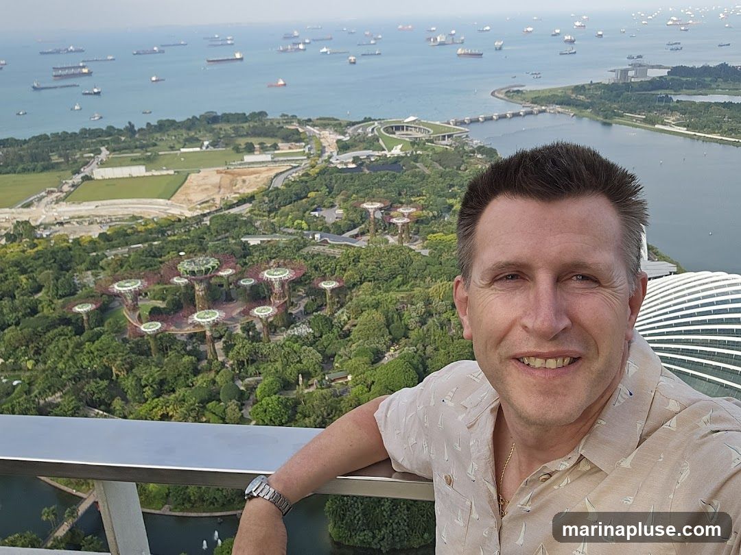 A man smiles at the camera, taking a selfie. He is leaning on a railing with a panoramic view of a city and a busy harbor filled with ships in the background. The cityscape includes lush green gardens with distinctive tree-like structures and modern architecture.