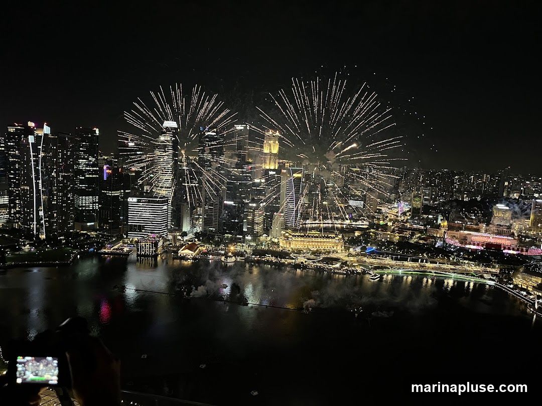 A wide-angle, nighttime aerial view captures a vibrant fireworks display illuminating a dense city skyline. Multiple bursts of fireworks, predominantly white and yellow, explode against the dark sky, reflecting on the calm water body in the foreground. The city is densely packed with illuminated skyscrapers and buildings, showcasing a sprawling urban landscape. In the lower left corner, a person is holding a camera, seemingly capturing the spectacle.