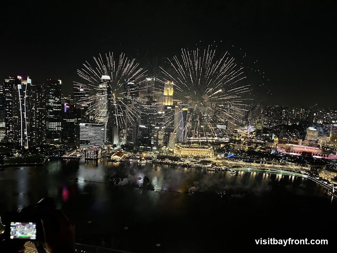 A wide-angle, nighttime aerial view captures a vibrant fireworks display illuminating a dense city skyline. Multiple bursts of fireworks, predominantly white and yellow, explode against the dark sky, reflecting on the calm water body in the foreground. The city is densely packed with illuminated skyscrapers and buildings, showcasing a sprawling urban landscape. In the lower left corner, a person is holding a camera, seemingly capturing the spectacle.