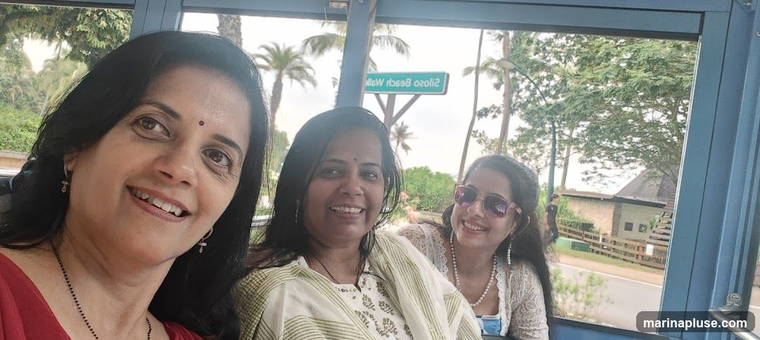 Three women are taking a selfie inside what appears to be a vehicle or tram. The woman on the left is in the foreground, smiling broadly. The other two women are behind her, also smiling. In the background, palm trees and lush greenery are visible, along with a sign that reads "Siloso Beach Walk". A building with a dark roof is also visible in the distance.