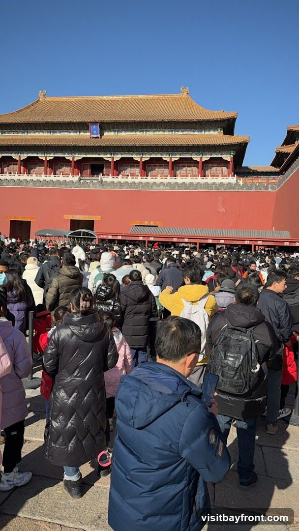 A large crowd of people are gathered in front of the Forbidden City under a clear blue sky. The traditional Chinese architecture of the palace complex is visible in the background, with its distinctive red walls and yellow tiled roofs. Many people are wearing winter clothing, suggesting a cool day. Some individuals are looking at their phones, while others are observing the surroundings. The scene depicts a popular tourist destination with a significant number of visitors.