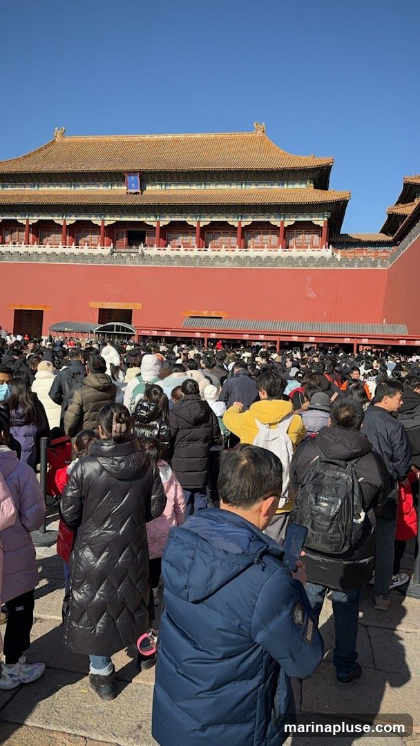 A large crowd of people are gathered in front of the Forbidden City under a clear blue sky. The traditional Chinese architecture of the palace complex is visible in the background, with its distinctive red walls and yellow tiled roofs. Many people are wearing winter clothing, suggesting a cool day. Some individuals are looking at their phones, while others are observing the surroundings. The scene depicts a popular tourist destination with a significant number of visitors.