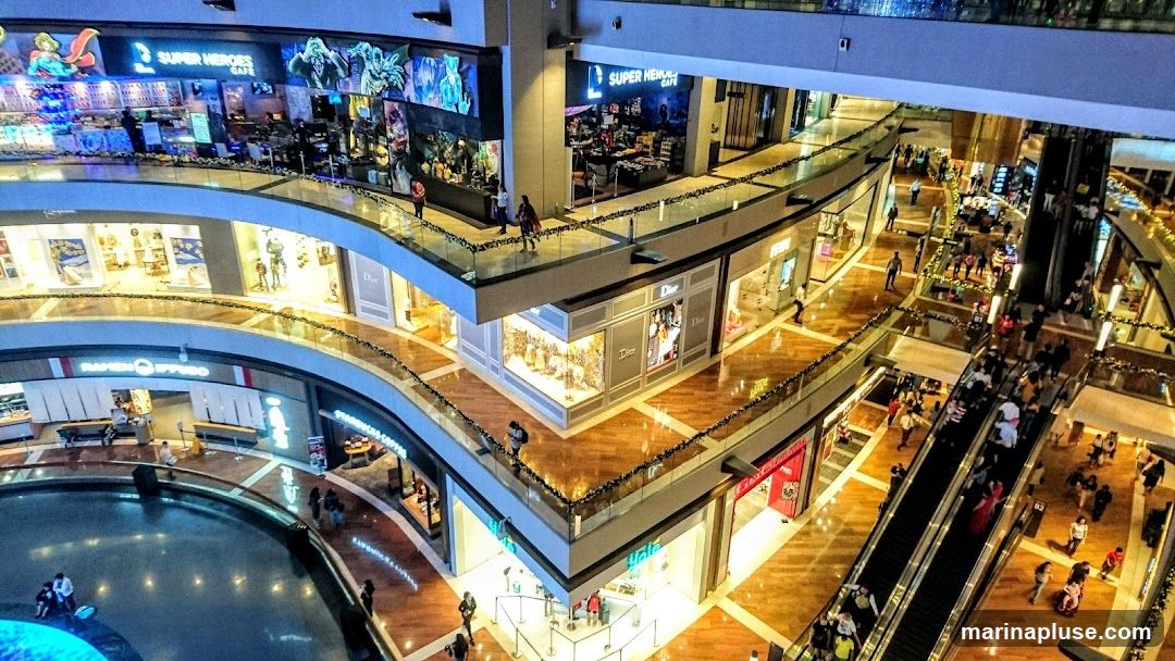 An aerial view of the interior of a modern shopping mall, showcasing multiple levels with shops, escalators, and shoppers. Prominent stores like Dior and Super Heroes Cafe are visible, along with other retail spaces and a central water feature.