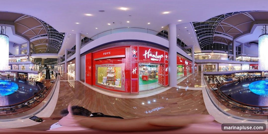 A panoramic view of a brightly lit, modern shopping mall interior. The focus is on the red facade of a Hamleys toy store, with its prominent logo and "TOYS" signage. The store features a display window showcasing plush toys. Surrounding the store are other shops and escalators. In the foreground, a person is visible holding a tablet. The mall has a large, circular water feature with blue lights and a glass railing overlooking it. The architecture includes a high, arched ceiling with skylights and large decorative chandeliers.