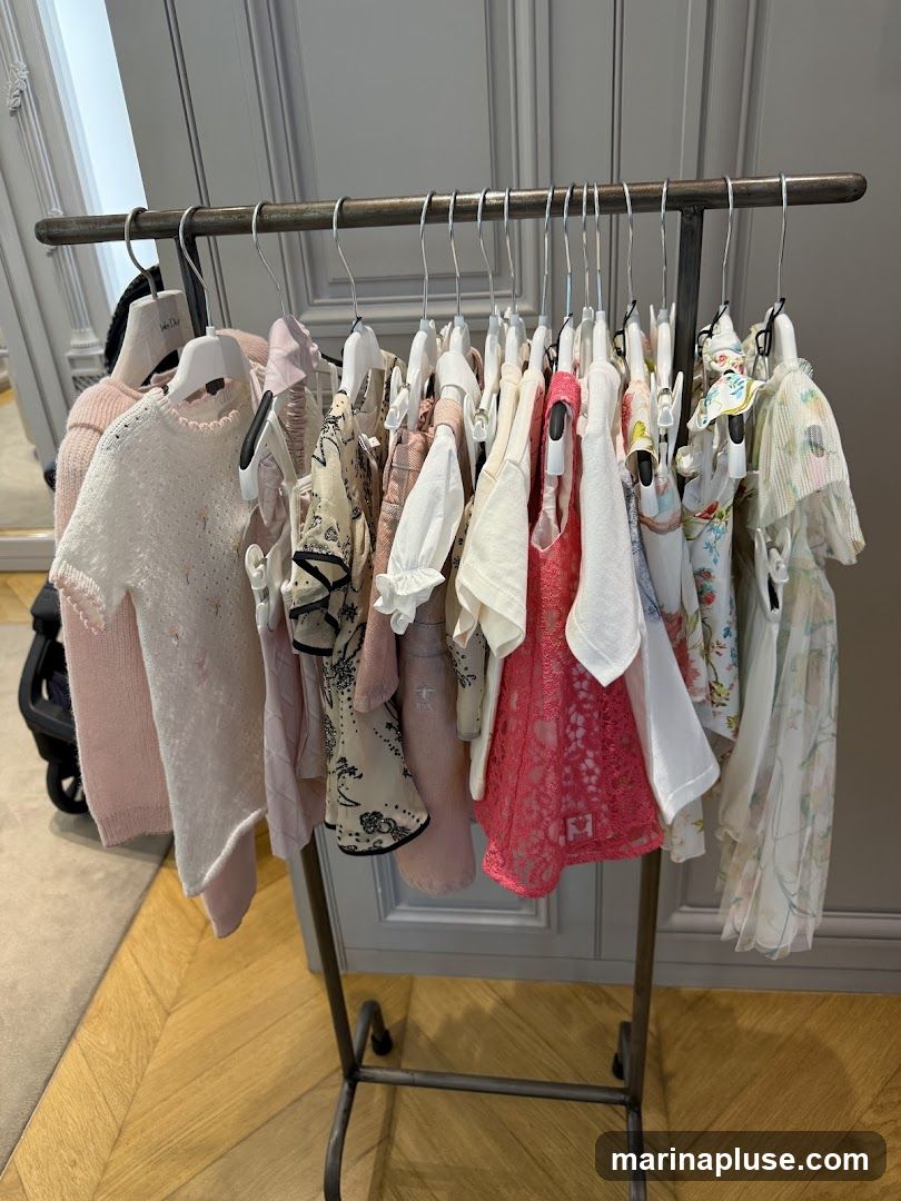 A metal clothing rack displays a variety of children's garments, including dresses, sweaters, and tops, in pastel colors and delicate fabrics. The clothes are hung on white hangers. The background shows a gray paneled wall and a wooden floor.