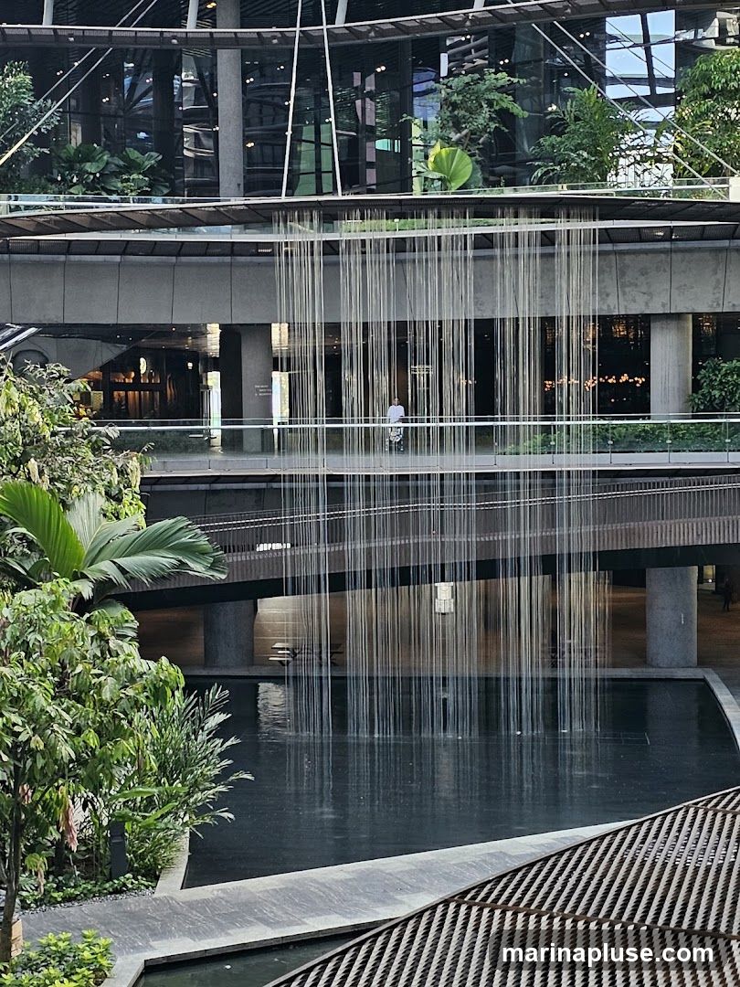 An interior view of a modern building with multiple levels, featuring a large indoor waterfall cascading into a dark pool. Lush green plants are visible on various balconies and levels, creating a natural and serene atmosphere. A person is seen on a walkway in the mid-ground, observing the waterfall. The architecture is contemporary with glass facades and concrete structures.