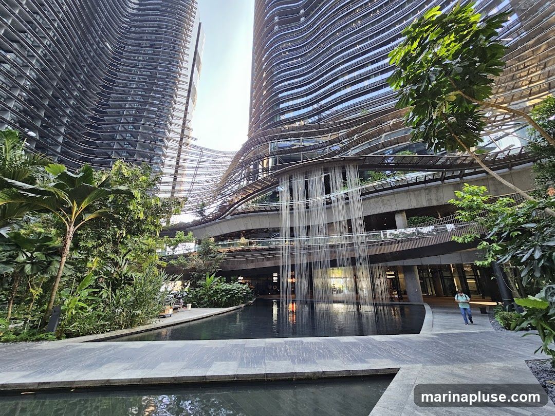 A wide-angle view of a contemporary architectural complex featuring two towering, wave-like buildings. A central atrium area includes a tranquil pool of water with a cascading waterfall feature. Lush greenery and trees are integrated throughout the space, creating a biophilic design. A person is walking on a pathway near the water. The overall impression is one of a modern, urban oasis.
