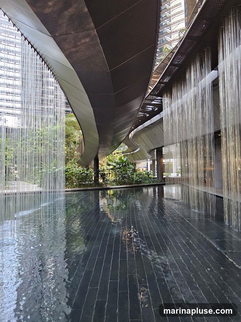 A contemporary architectural space featuring a shallow pool of water with dark tiled flooring. Above the pool, a curved, multi-layered structure with metallic and fabric elements creates a dramatic canopy. Vertical strands of what appear to be glass or metal hang from the structure, resembling a curtain of water or light. Lush greenery is visible in the background, suggesting an indoor garden or atrium setting within a modern building. The reflections in the water add depth and visual interest to the scene.