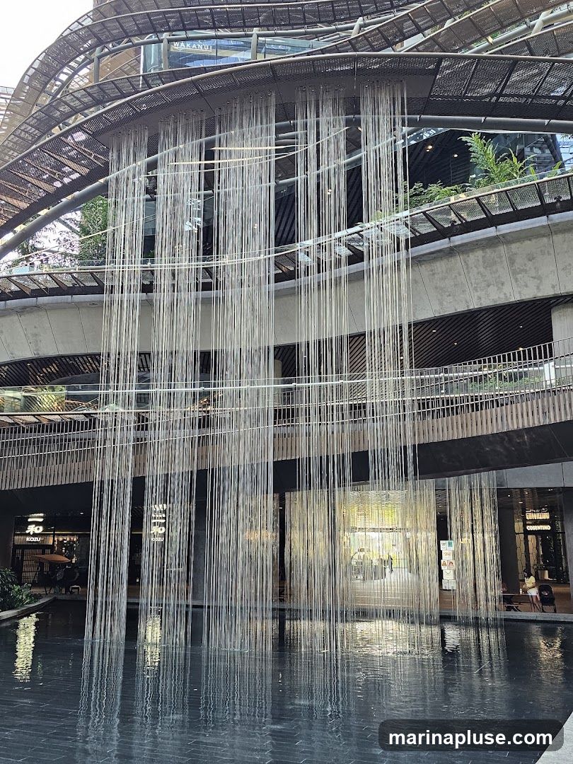 A modern indoor space with a multi-level design featuring a large waterfall installation made of numerous thin strands. The strands cascade from an overhead structure into a dark, reflective pool at the bottom. The background shows various shops and walkways on different levels, with some greenery visible.