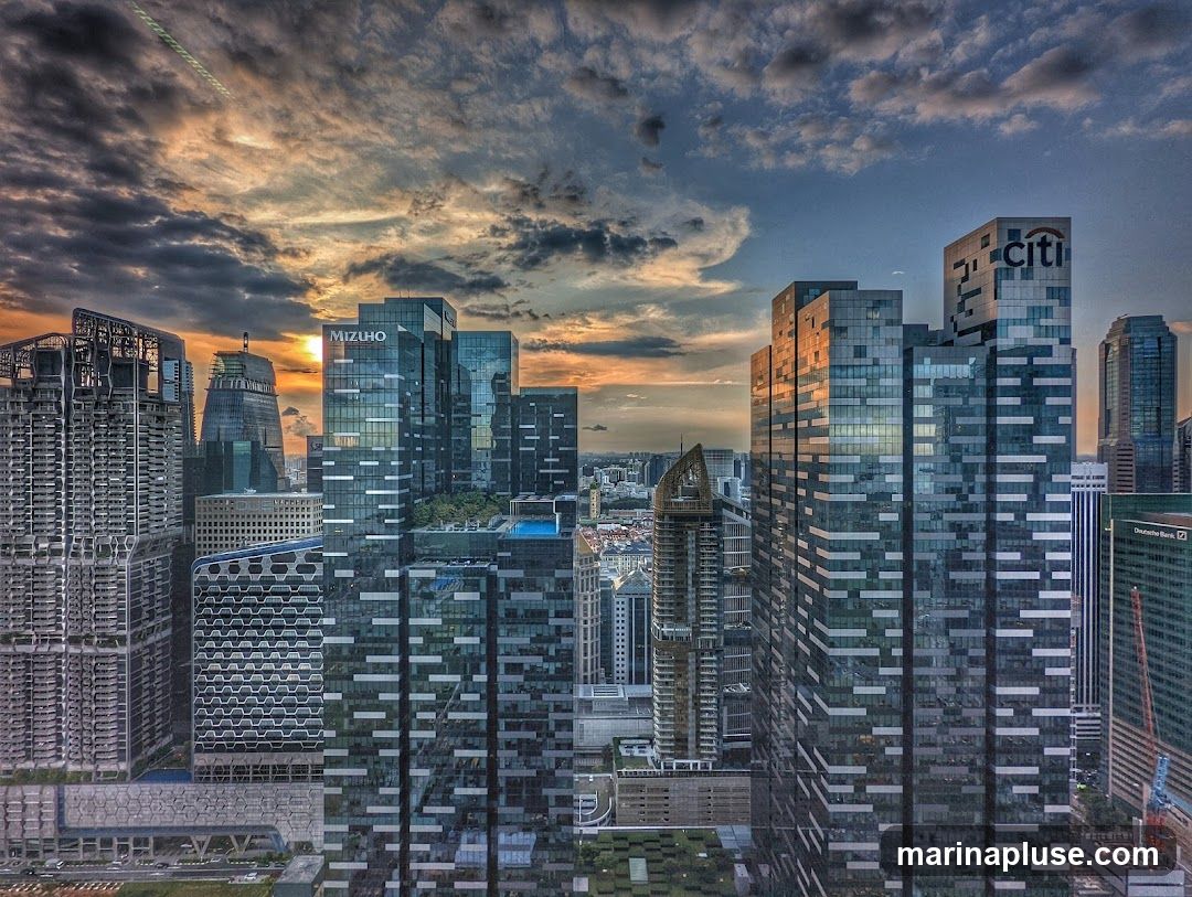 A panoramic view of a modern cityscape at sunset, with towering skyscrapers reflecting the warm hues of the sky. Clouds are scattered across the sky, with the sun setting behind some of the buildings. Prominent building names like 'CITI' and 'MIZUHO' are visible.