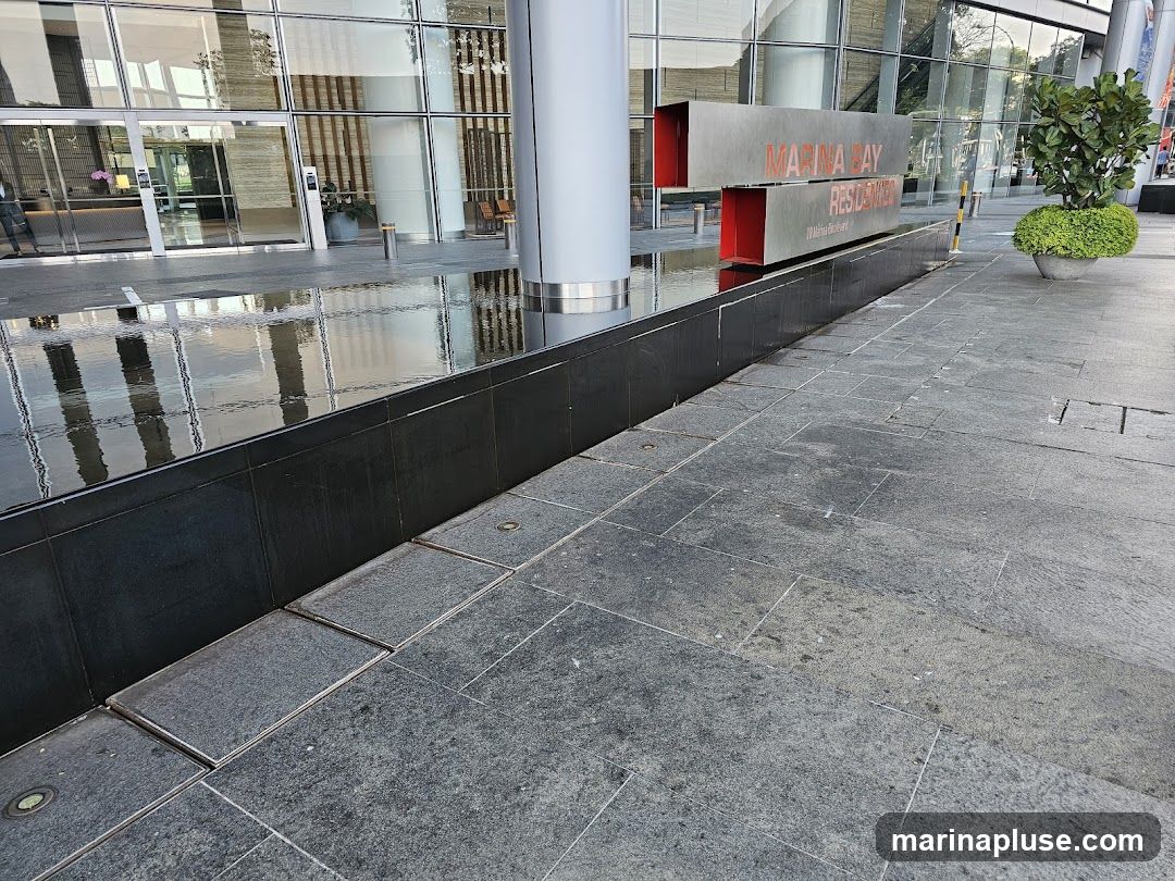 The exterior of the Marina Bay Residences building, featuring a modern facade with large glass windows and a prominent metal sign. A water feature runs along the front of the building, reflecting the architecture. Paved walkways and a potted plant complete the scene.