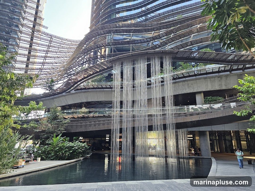 A wide shot of a contemporary building with a unique, undulating facade. A man-made waterfall cascades down from an elevated walkway, creating a striking visual effect. Lush greenery is integrated into the architecture, with trees and plants visible on various levels. A reflective pool in the foreground mirrors the structure and the falling water. Several people are seen walking around the area, suggesting it is a public space or shopping complex.