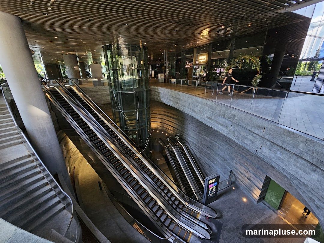 A wide shot of a modern atrium with multiple escalators and staircases. A glass elevator shaft is visible in the center. People are seen walking on an upper level walkway, and a decorative floral hoop is displayed. The architecture features concrete and glass elements with a slatted ceiling.