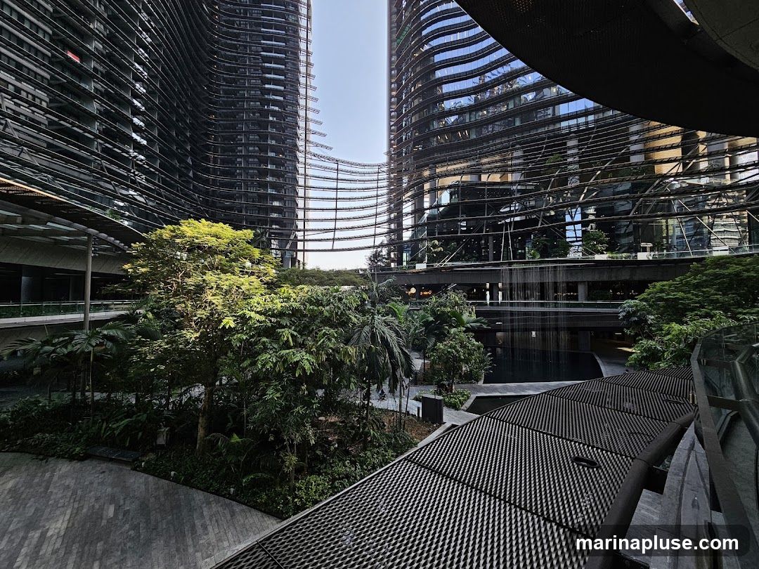 A view of a modern architectural complex featuring two tall, curved buildings with reflective glass facades. In the foreground and between the buildings, there is a lush green garden with various trees and plants. A water feature cascades down from an elevated walkway, creating a serene atmosphere. The ground level appears to be paved with a patterned stone.