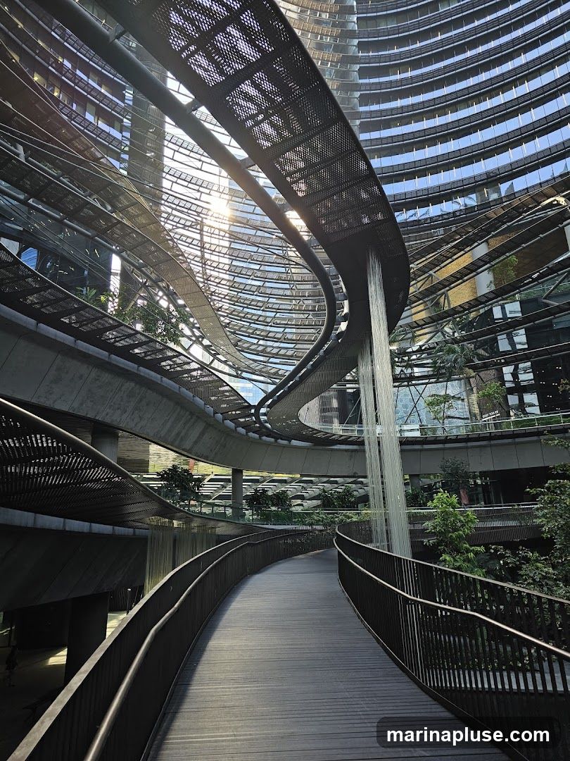 A curved wooden walkway with metal railings winds through a multi-level modern architectural space. The structure features curved glass facades, metal grating walkways, and lush greenery. A thin waterfall cascades down from an upper level, adding a natural element to the urban design. Sunlight streams through the open roof, illuminating the scene.