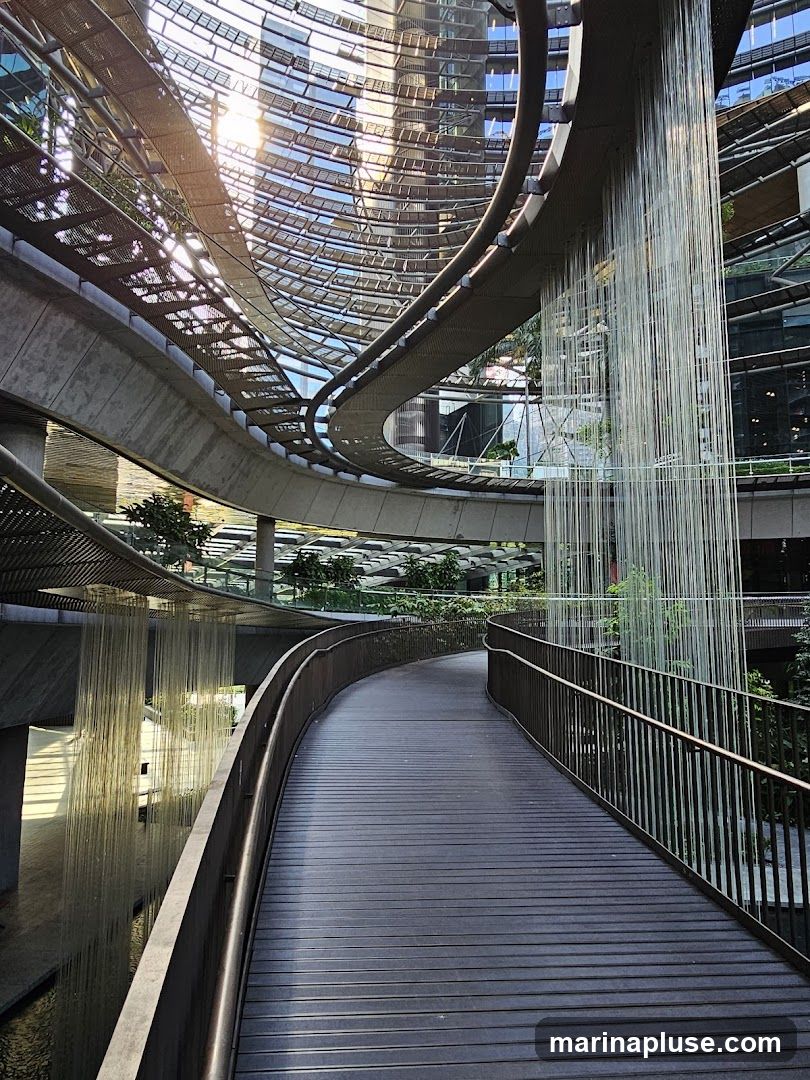 A wooden walkway curves through a modern architectural space with a glass roof and surrounding buildings. Water cascades down from an elevated structure, creating a curtain-like effect. Sunlight streams through the glass, casting shadows and reflections on the walkway and water. Lush greenery is visible in the background, integrated into the multi-level design.