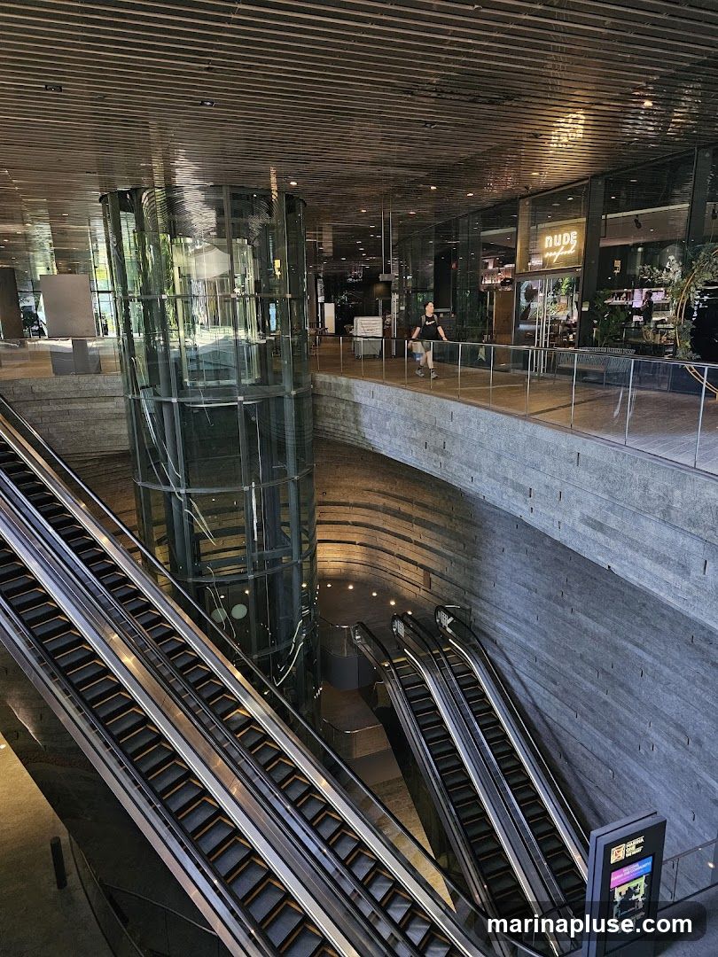 A wide shot of a modern interior space featuring multiple escalators leading up and down. A glass elevator shaft is visible in the center. A walkway overlooks the escalators, with several shops and restaurants visible in the background. One shop has a sign that reads "NUDE seafood". A person is walking on the walkway.