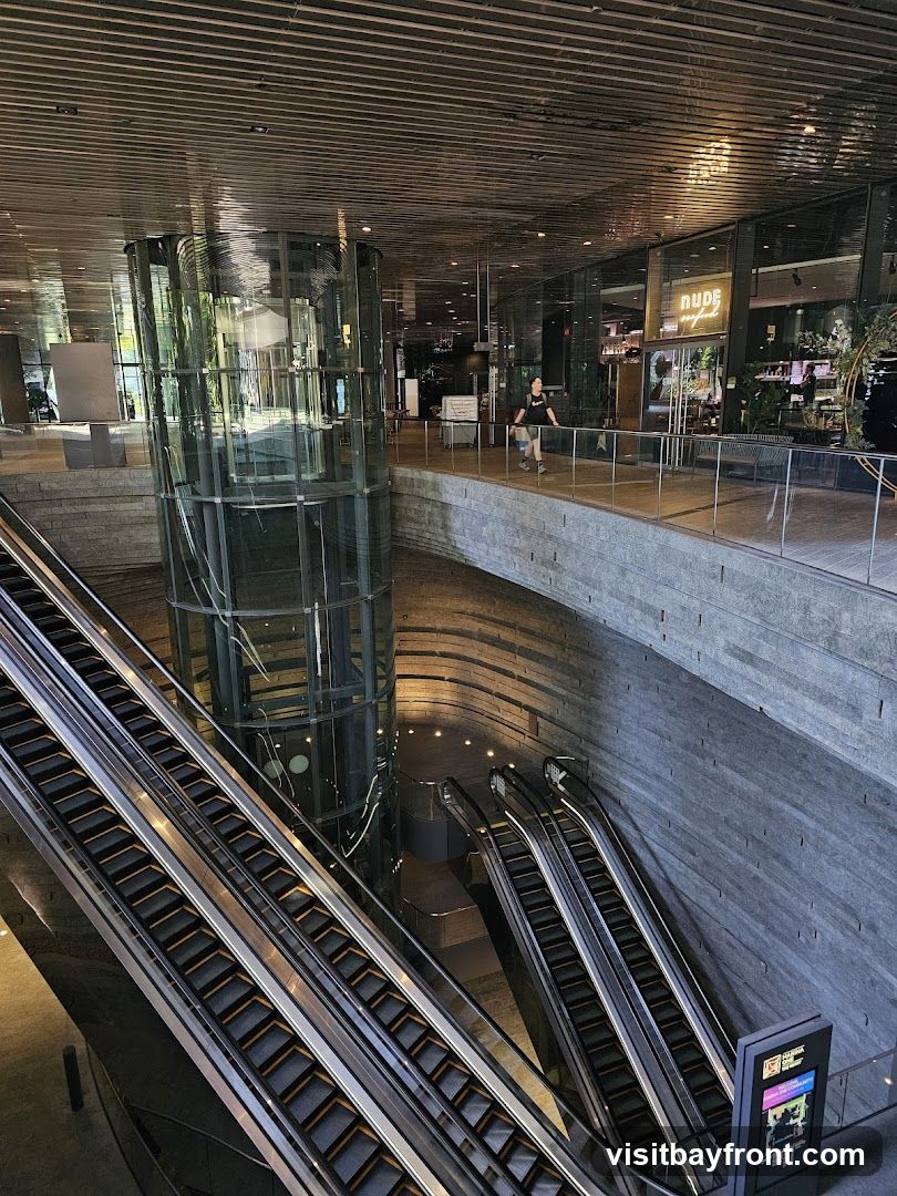 A wide shot of a modern interior space featuring multiple escalators leading up and down. A glass elevator shaft is visible in the center. A walkway overlooks the escalators, with several shops and restaurants visible in the background. One shop has a sign that reads "NUDE seafood". A person is walking on the walkway.