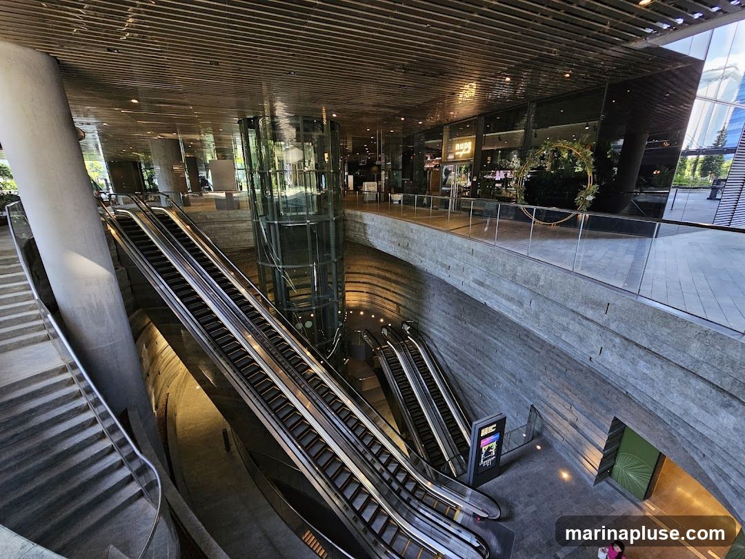 An interior view of a modern building atrium featuring multiple escalators, a glass elevator shaft, and a contemporary architectural design with concrete and metallic elements. The upper level shows storefronts with large glass windows, reflecting the exterior. A decorative circular floral arrangement is visible on the upper walkway.