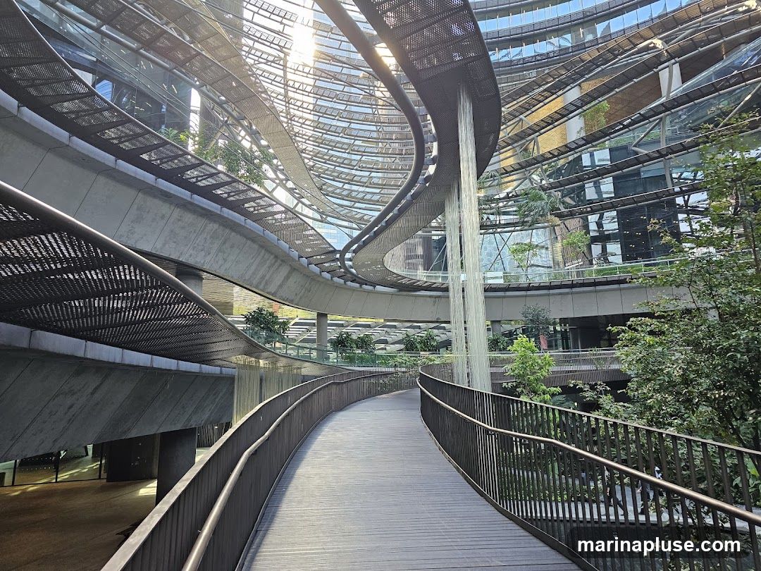 A curved wooden walkway with metal railings winds through a multi-level indoor space. Above, a complex architectural structure of glass and metal forms a ceiling, with sunlight filtering through. A waterfall cascades down from an upper level. Lush greenery and trees are integrated into the design, creating a serene, modern garden environment within a building.