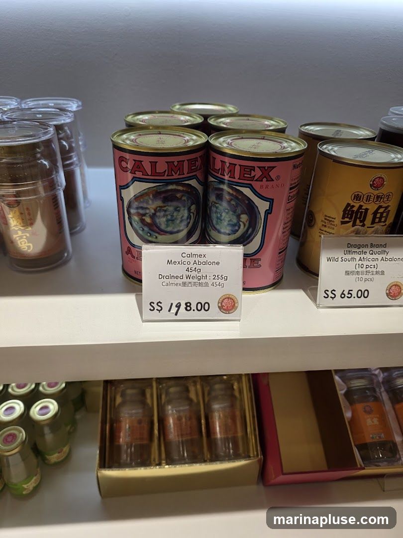 A shelf displaying various canned abalone products. In the foreground, there are cans of Calmex Mexico Abalone and Dragon Brand Wild South African Abalone. Below, there are gift boxes containing small bottles, possibly bird's nest soup.
