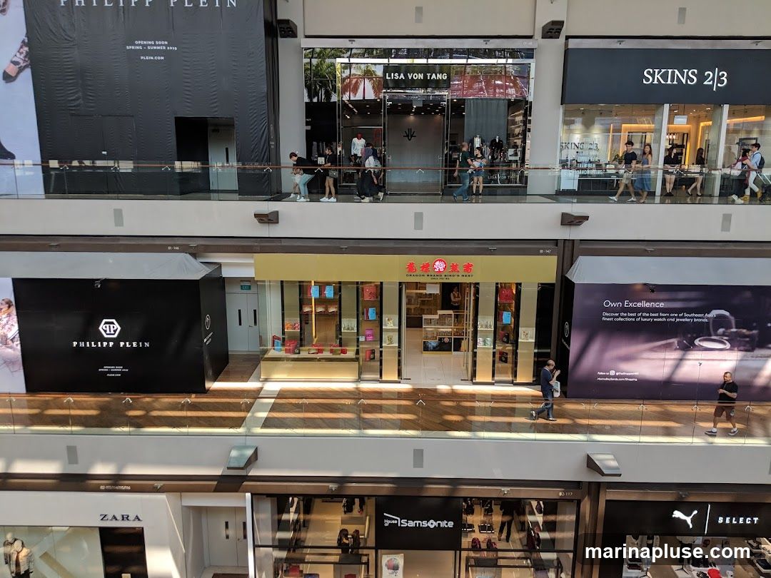 An interior view of a multi-level shopping mall with various storefronts visible. On the upper level, storefronts for PHILIPP PLEIN, LISA VON TANG, and SKINS 2/3 are seen. On the middle level, a PHILIPP PLEIN advertisement and a storefront for DRAGON BRAND BIRD'S NEST are visible. On the lower level, storefronts for ZARA, Samsonite, and Puma are present. People are seen walking on the elevated walkways.
